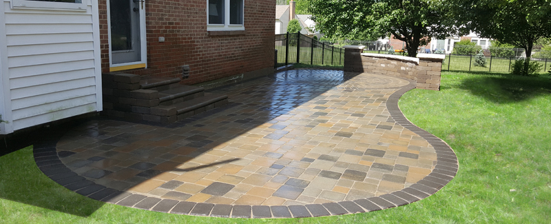 A paver patio in a back yard with table and chairs and surrounding a pool on a summer day.